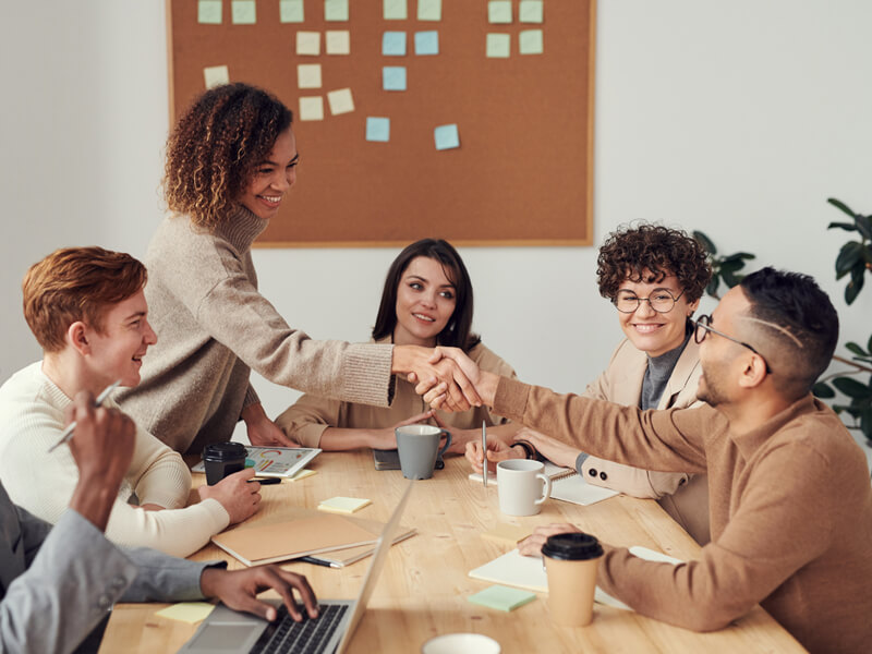 Eine Gruppe von 6 Menschen ist an einem Tisch zu sehen. Eine Person davon steht und schüttelt die Hand einer am Tisch seitzenden Person